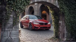 A photo of a red BMW M3 parked on a cobblestone street. The car is in the background, with its sleek lines and distinctive kidneys grille illuminated. The foreground is a stone archway, with ivy growing over it. The background contains a building with multiple arches. The lighting is warm.