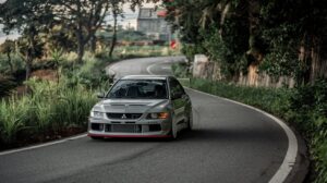 A photo of a street-tuned Mitsubishi Lancer Evo VIII driving along a winding country road. The road is surrounded by lush greenery, with trees and plants on both sides. The car is moving at a moderate speed, with its tires grazed along the edge of the road. The car's body has been modified with a rear wing and a front lip spoiler. The background contains a few buildings in the distance.