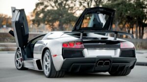 A photo of a Lamborghini Murcielago SV with the rear end and both doors open. The car is parked outdoors. The background contains a few trees. The lighting is bright.