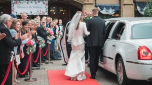 A photo of a bride and groom exiting a white limousine. The bride is wearing a white dress and a veil, while the groom is wearing a tuxedo. They are walking down a red carpet. There is a crowd of people behind a rope, clapping and cheering. There are also flowers and a banner with the text "Welcome to the Wedding".