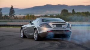 A rear photo of a metallic silver Mercedes SLR McLaren doing a burnout on an empty track. The car is static, with smoke billowing from the rear tires. The background is a scenic landscape with mountains, trees, and a clear sky.